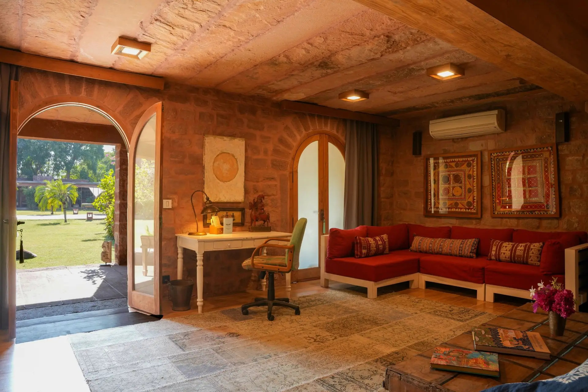 A sandstone sitting room with scalloped arches opening onto the garden, a red sofa and framed Rajasthani textiles